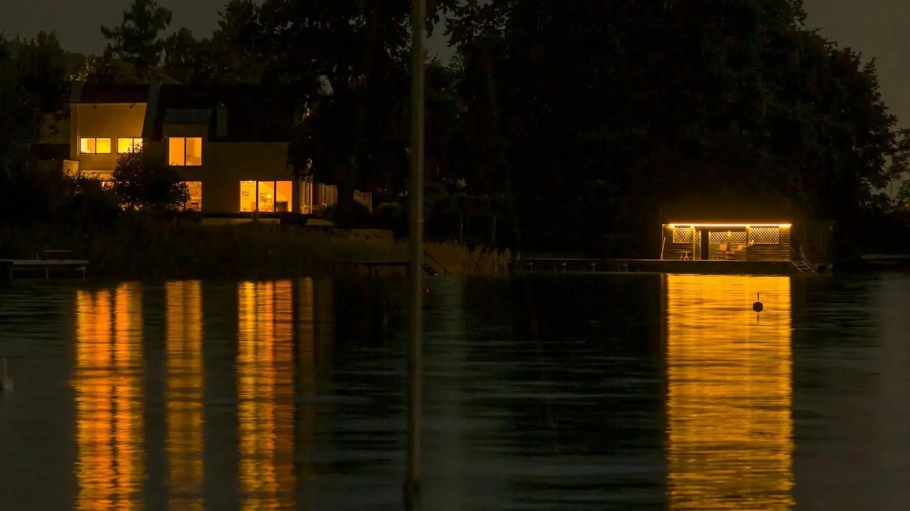 A still boat in the middle of a lake; sunset-like house lights in the backdrop.